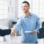 Young people in handshake at modern office. One man and two women in formalwear, man holding papers. Monitor, documents, phone and tools on desk. Windows, copy machine and a bike on background.