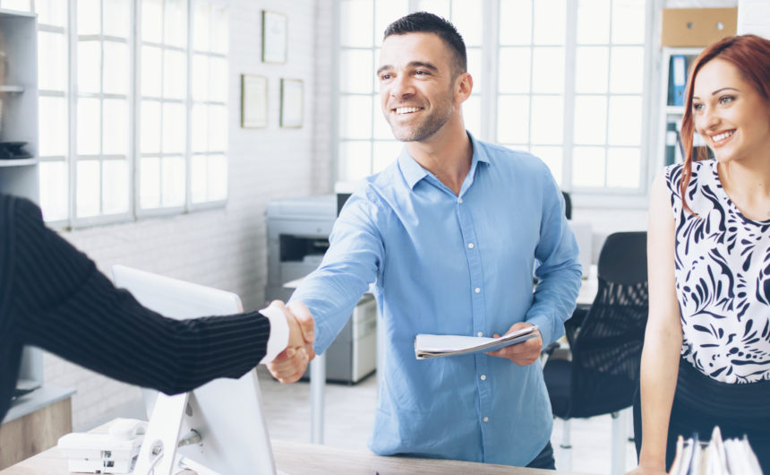 Young people in handshake at modern office. One man and two women in formalwear, man holding papers. Monitor, documents, phone and tools on desk. Windows, copy machine and a bike on background.