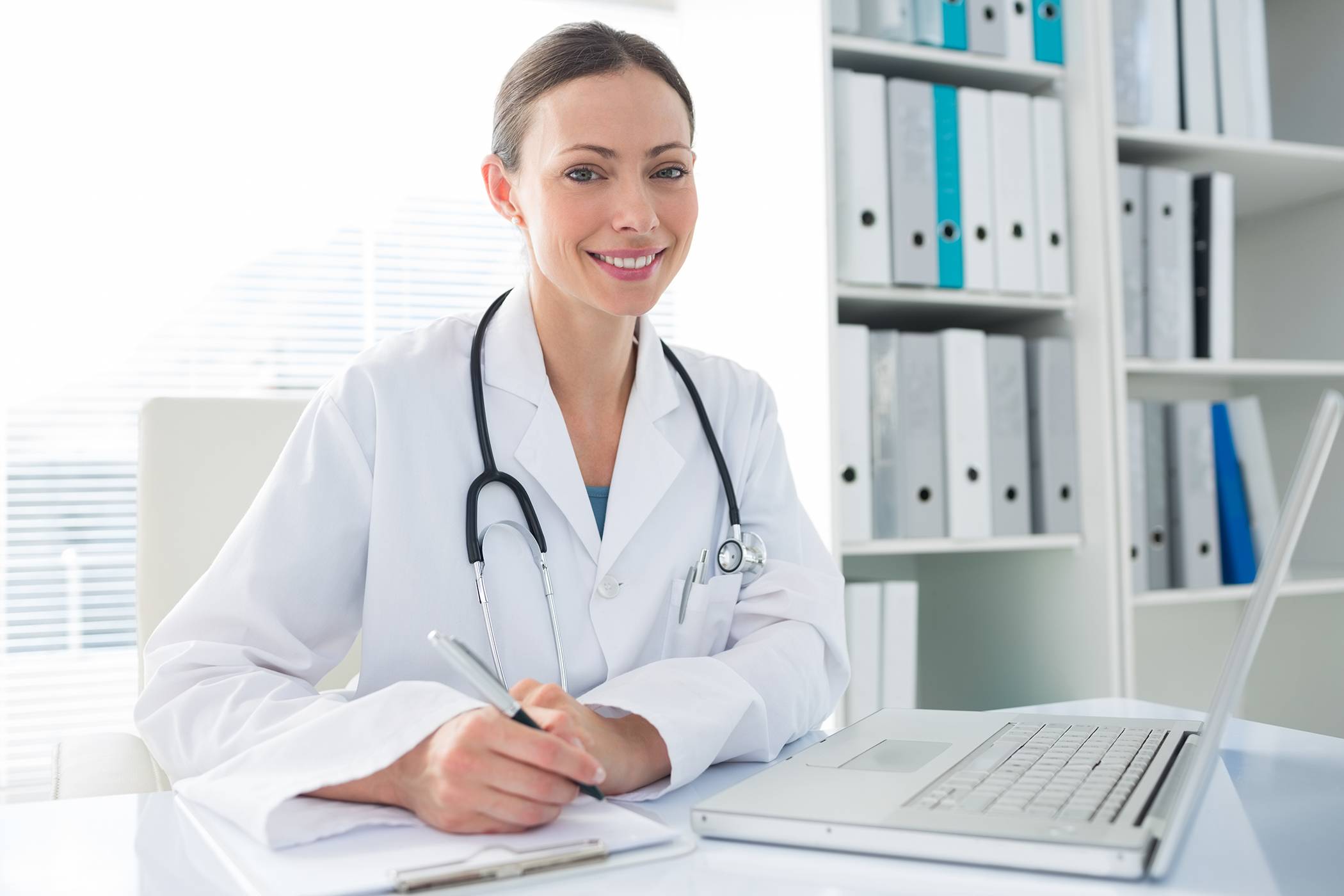 Beautiful female doctor with laptop and clipboard sitting at desk in clinic